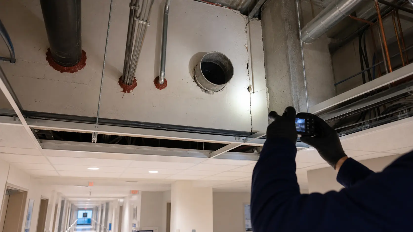 Inspector documenting an abandoned sleeve and unsealed joint above a hospital ceiling